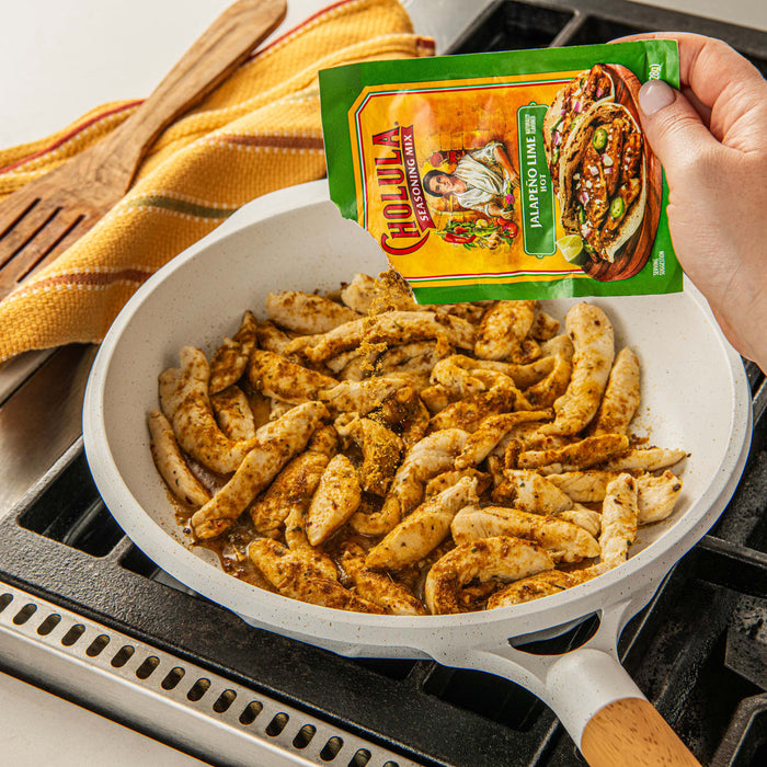 A hand sprinkles Cholula Jalapeno Lime seasoning on chicken strips in a white skillet.