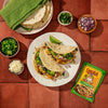 A plate of tacos with jalapeno, red onion, cilantro, and cotija cheese, with a packet of Cholula Jalapeno Lime Hot seasoning mix.
