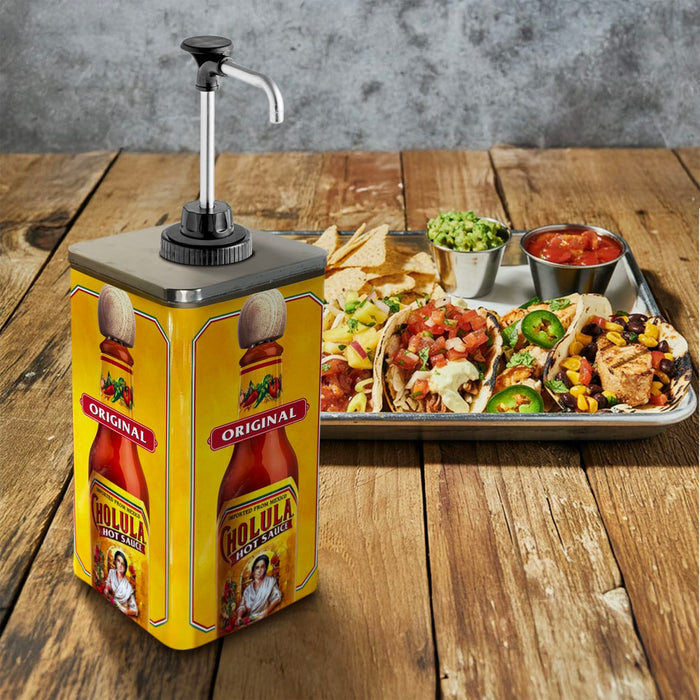 A large yellow pump dispenser of Cholula Original Hot Sauce sits on a wooden table next to a tray of loaded tacos and dipping salsas.