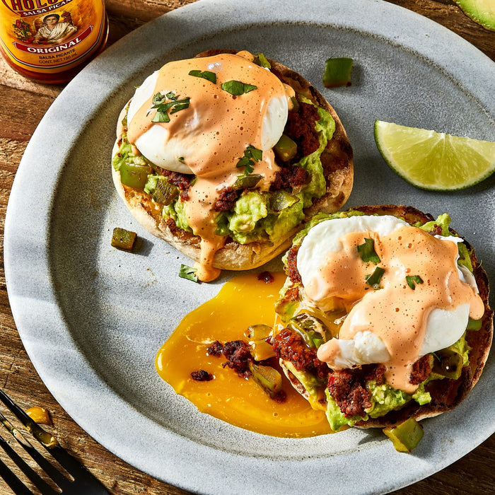 A bottle of Cholula Original Hot Sauce sits next to a plate of Mexican style eggs benedict featuring poached eggs topped with creamy chipotle sauce and chorizo on toasted English muffins.
