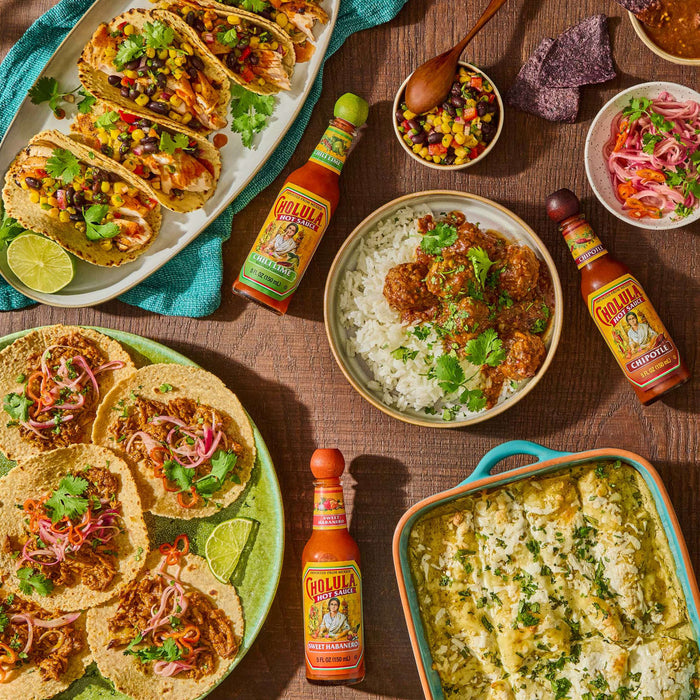A table with a variety of Mexican dishes, including tacos, enchiladas, and rice with meatballs, all topped with Cholula hot sauce.