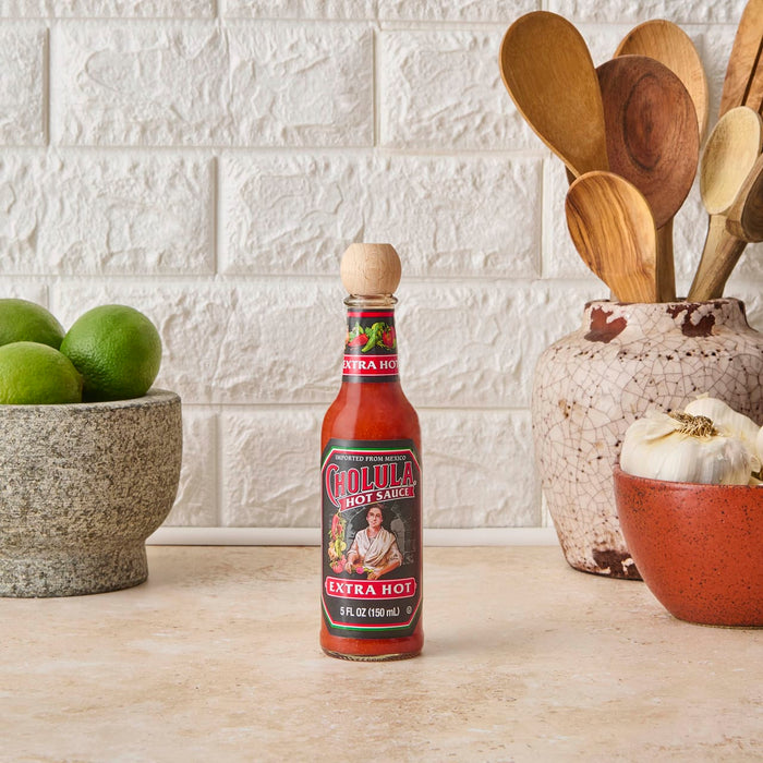 A bottle of Cholula Extra Hot Hot Sauce sits on a countertop with a bowl of limes and a bowl of garlic.