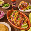 A plate of fish tacos with red cabbage slaw, cilantro, and lime wedges, with a bottle of Cholula Hot Sauce in the background.