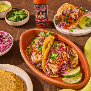 A plate of fish tacos with red cabbage slaw, cilantro, and lime wedges, with a bottle of Cholula Hot Sauce in the background.