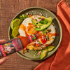A hand is pouring Cholula Hot Sauce over a plate of eggs, avocado, and tortilla chips.
