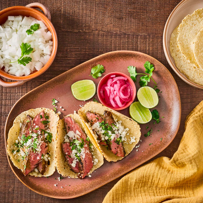 Three tacos with steak, onions, cilantro, and cheese on a plate with limes, pickled onions, and cilantro.