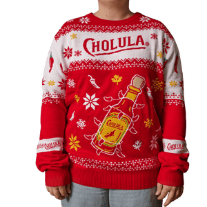 A person wears a festive red and white holiday sweater featuring the Cholula logo and a large illustration of a hot sauce bottle wrapped in lights.