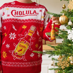 A person wearing a festive red and white holiday sweater featuring the Cholula logo hangs a miniature bottle ornament on a decorated Christmas tree.