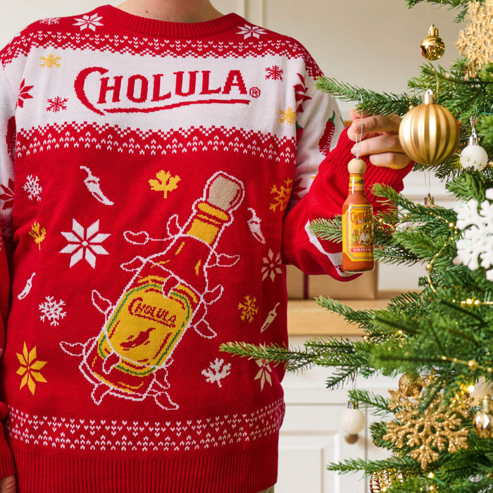 A person wearing a festive red and white holiday sweater featuring the Cholula logo hangs a miniature bottle ornament on a decorated Christmas tree.