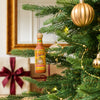 A festive holiday scene features a miniature bottle of Cholula Hot Sauce Original hanging as a unique ornament on a decorated Christmas tree.