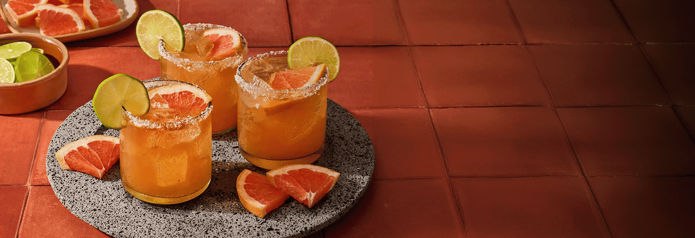 Three glasses of orange juice with lime wedges and grapefruit slices on a stone coaster.