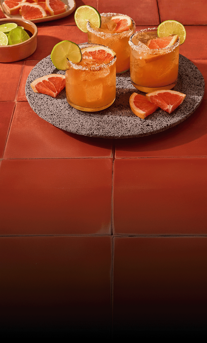 Two orange cocktails with lime wedges and grapefruit slices on a textured stone coaster.