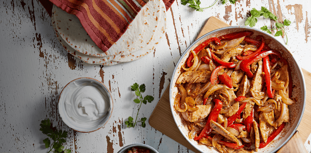 Fajita dish with chicken and bell peppers on a wooden table with tortillas and sour cream.