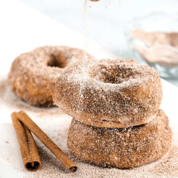 Two cinnamon sugar donuts are stacked on top of each other with a third donut in the background, with cinnamon sticks and powdered sugar sprinkled on top.