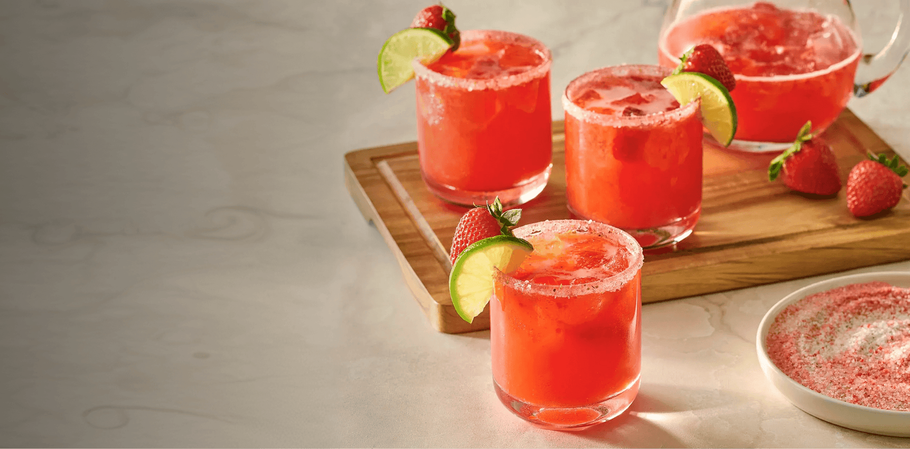 Four glasses of red cocktails with lime wedges and strawberries on a wooden board.