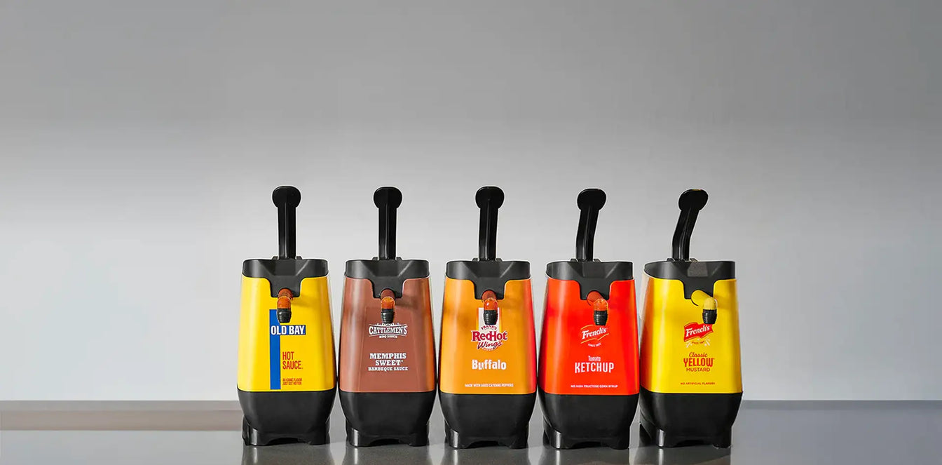 Five condiment dispensers on a reflective surface with a gray background