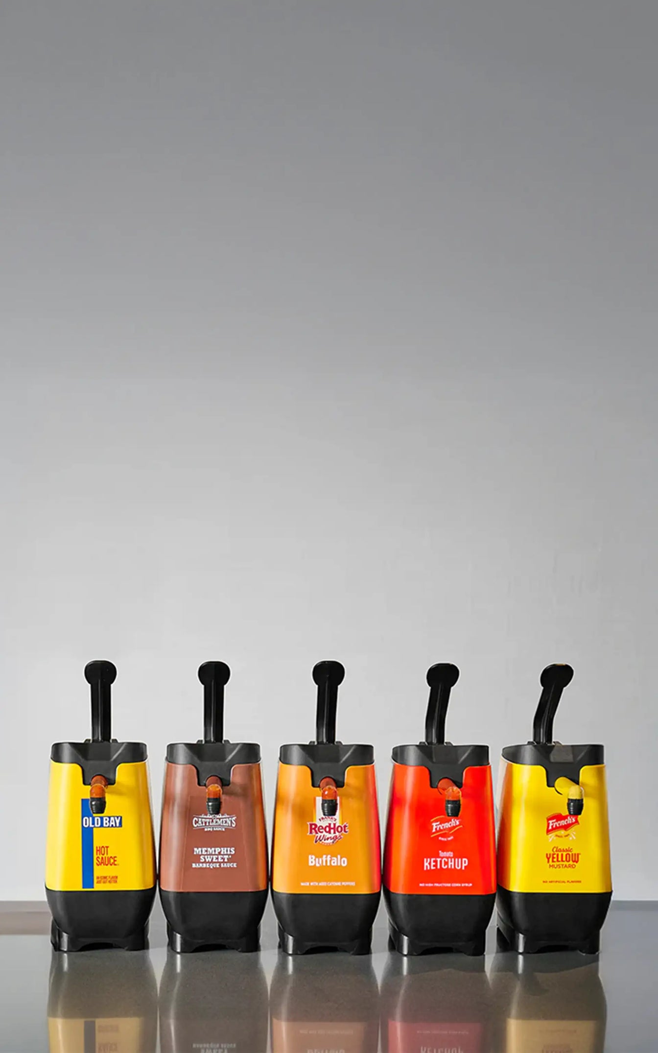 Five condiment dispensers on a reflective surface with a gray background