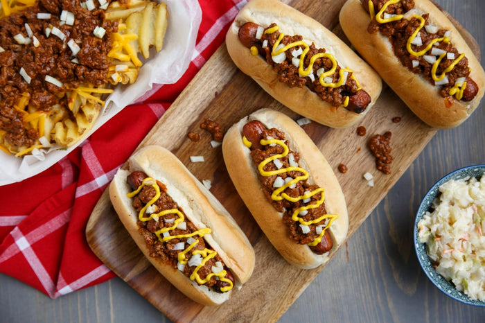 Four hot dogs topped with chili, mustard, and onions are on a wooden cutting board next to a bowl of coleslaw.