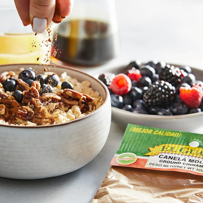 A hand sprinkles cinnamon on a bowl of oatmeal with blueberries and pecans, next to a bowl of berries and a bag of El Guapo ground cinnamon.