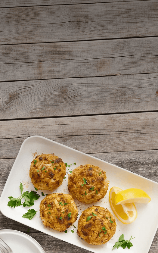 Four crab cakes on a white plate with lemon wedges and parsley on a wooden surface