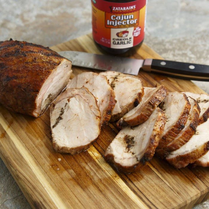 A sliced pork loin injected with Zatarains Cajun Injector Creole Garlic marinade sits on a wooden cutting board with a knife.
