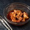 A glass bowl filled with marinated meat and sesame seeds sits next to a pair of tongs on a dark surface.