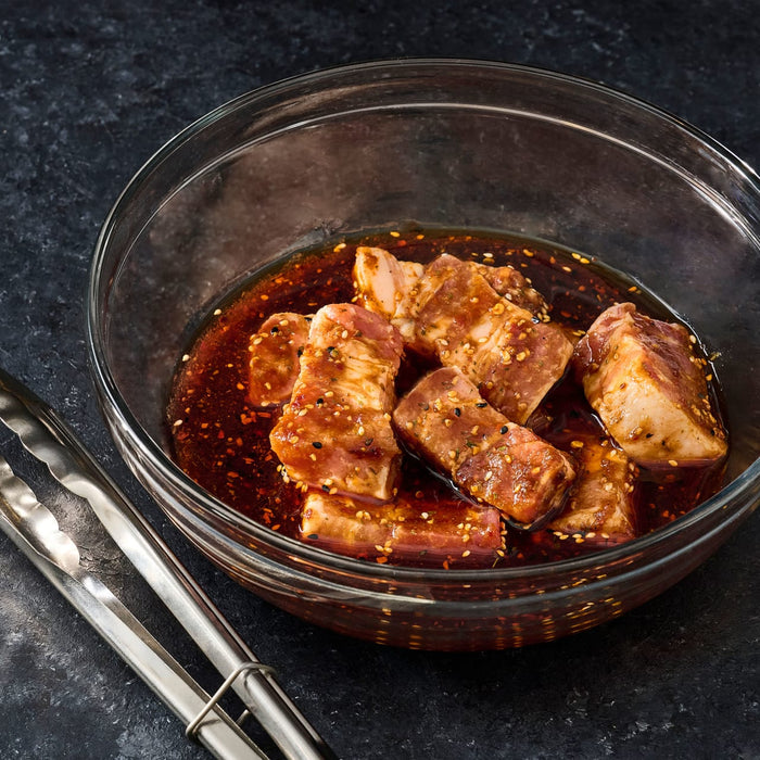 A glass bowl filled with marinated meat and sesame seeds sits next to a pair of tongs on a dark surface.