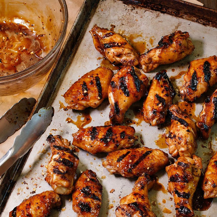 A tray of grilled chicken wings with a bowl of sauce and tongs.