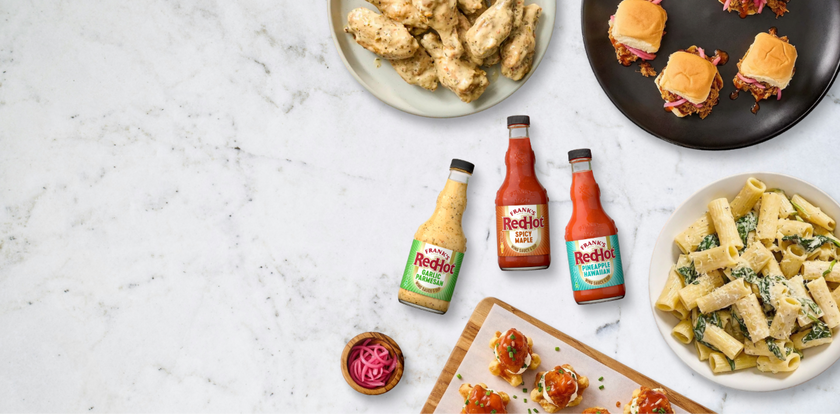 Three bottles of Frank's RedHot hot sauce on a marble surface with plates of food including sandwiches and pasta.