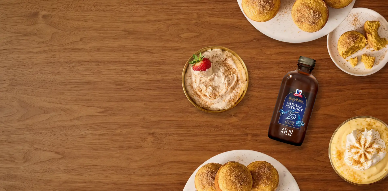 A bottle of vanilla extract placed on a wooden tabletop, surrounded by plated pastries, a dessert with a strawberry, and a yellow drink.