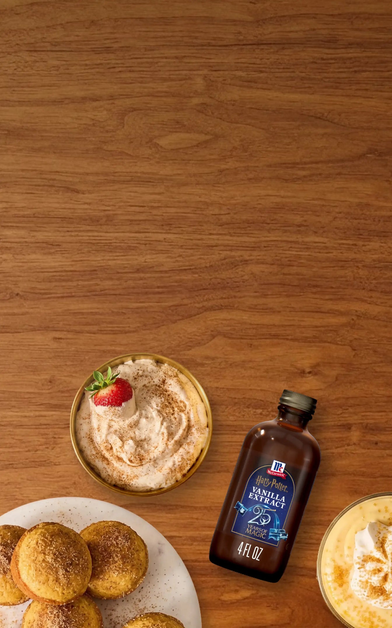 A bottle of vanilla extract placed on a wooden tabletop, surrounded by plated pastries, a dessert with a strawberry, and a yellow drink.