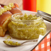 A jar of green relish sits on a white plate next to a hot dog bun and a spoon.