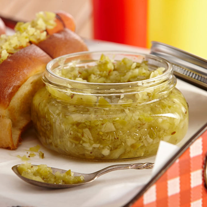 A jar of green relish sits on a white plate next to a hot dog bun and a spoon.