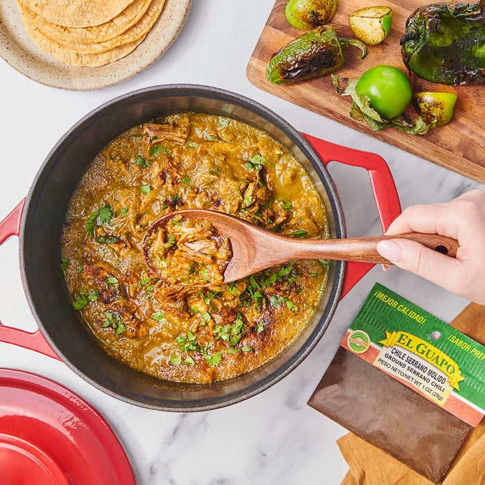 A pot of El Guapo Chile Serrano Molido Ground Serrano Chili with a wooden spoon and a bag of El Guapo Chile Serrano Molido Ground Serrano Chili.