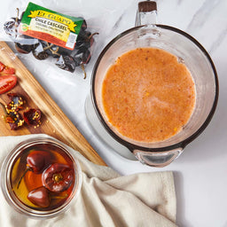 A close-up of a blender filled with a red sauce, a bowl of red peppers, and a bag of El Guapo Chile Cascabel peppers.