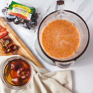 A close-up of a blender filled with a red sauce, a bowl of red peppers, and a bag of El Guapo Chile Cascabel peppers.