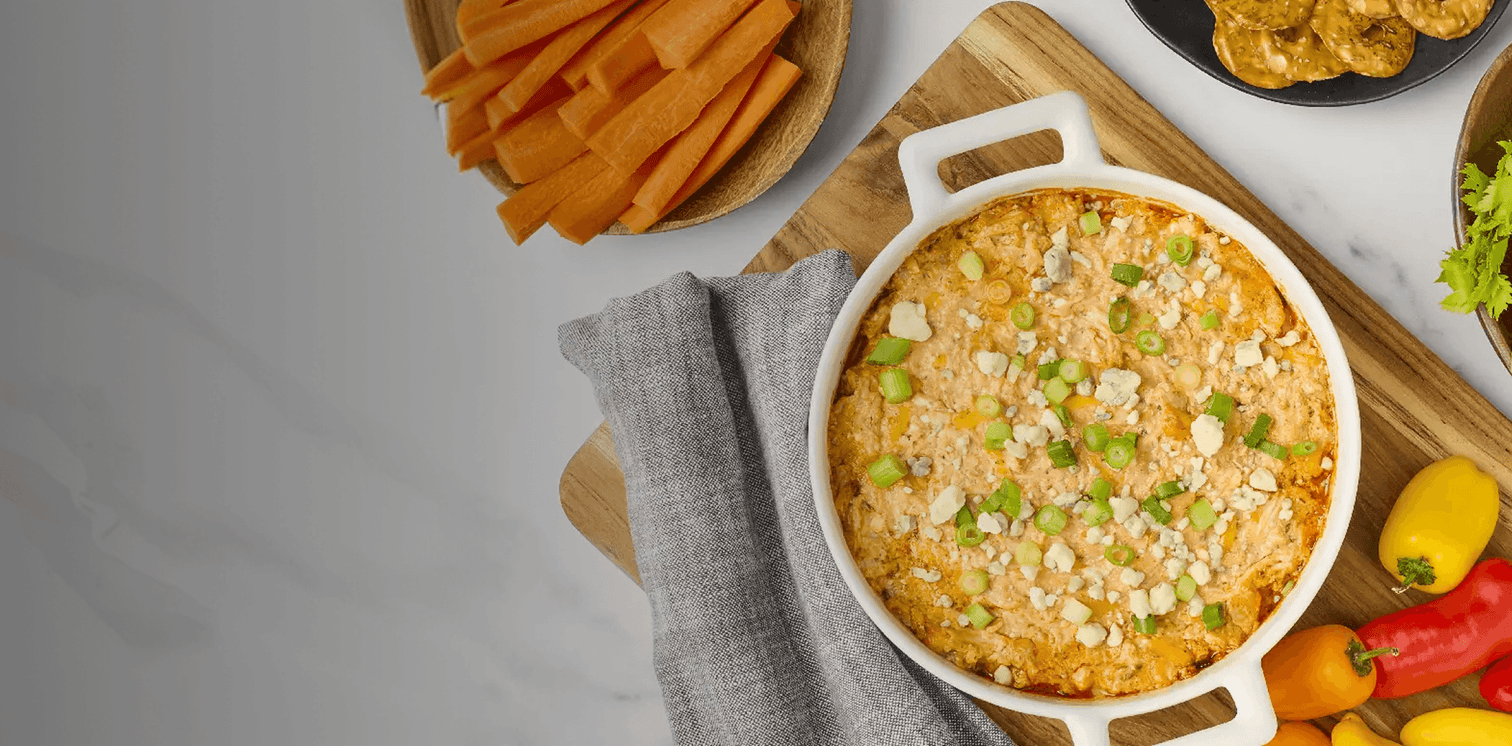 Baked dip with vegetables in a white dish on a wooden board, surrounded by chips and vegetables.