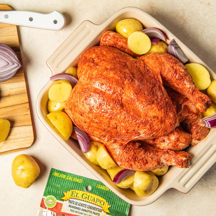 A close-up of a seasoned chicken with potatoes and onions in a baking dish, with a package of El Guapo Seasoned Annatto Paste in the foreground.
