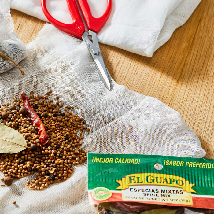 A close-up of a bag of El Guapo spices with a red chili pepper, coriander seeds, and bay leaves.