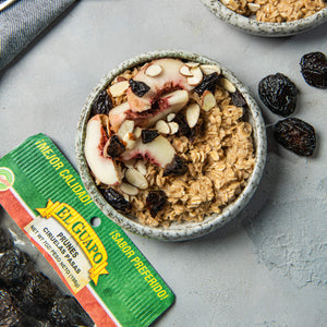 A bowl of oatmeal with peaches, almonds, and prunes, with a package of El Guapo prunes in the foreground.