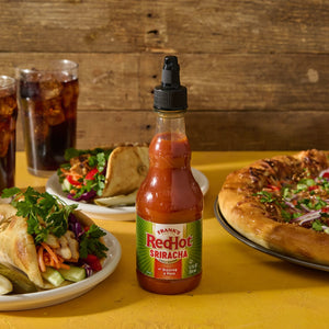 A bottle of Franks RedHot Sriracha chili sauce sits on a yellow table with a plate of food in front of it.