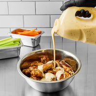 Hand pouring creamy sauce over fried chicken in a metal bowl with celery and carrots on the side.