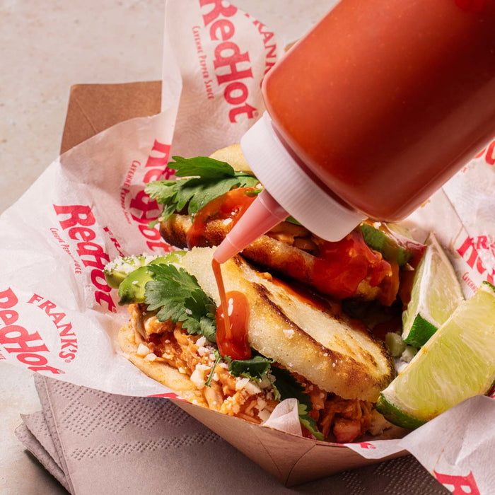 A RedHot sauce bottle pouring sauce on a arepa with chicken, avocado, cilantro and lime wedges.