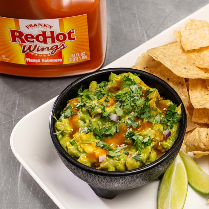 A bowl of guacamole with Franks RedHot Mango Habanero sauce, lime wedges, and tortilla chips.