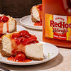 A slice of cheesecake topped with strawberry sauce and a bottle of Franks RedHot Mango Habanero sauce.