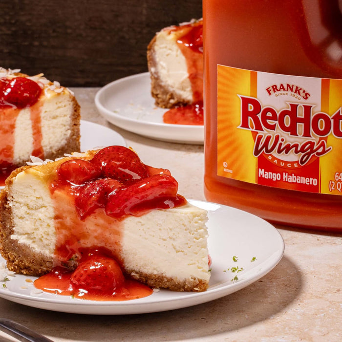 A slice of cheesecake topped with strawberry sauce and a bottle of Franks RedHot Mango Habanero sauce.