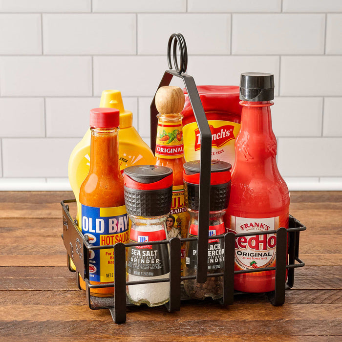 A black metal condiment caddy holds a variety of flavor enhancers including Old Bay Hot Sauce, McCormick Sea Salt Grinder, McCormick Black Peppercorn Grinder, Frank s RedHot Original Cayenne Pepper Sauce, Cholula Hot Sauce, and French s Mustard.