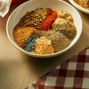 A white bowl filled with various whole and ground spices including coriander seeds, fennel, chili powder, and dried herbs is arranged on a wooden surface next to a red checkered napkin.