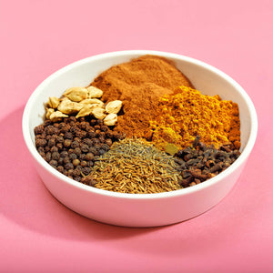 A white bowl on a pink background contains a variety of whole and ground spices including black peppercorns, cardamom pods, cumin seeds, cloves, turmeric, and cinnamon.