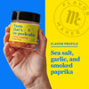 A hand holds a jar of Flavor Makers im-peck-able chicken topping seasoning, with a yellow background showing the Flavor Maker logo and the flavor profile.